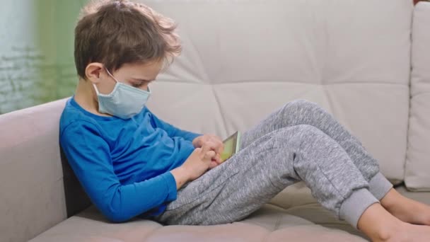 Un băiețel arătos în fața portretului camerei care stă pe canapea concentrat jucând jocul pe smartphone în timp ce este în carantină copilul care poartă masca de protecție — Videoclip de stoc