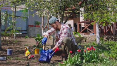 Bahçede bir adam bir su bidonuna su koyar ve çiçekleri sular güneşli bir günde arka planda bir kadın musluğu açar.