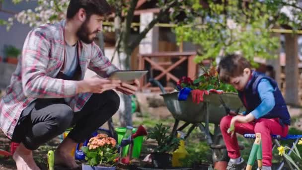 Tiempo feliz hijo y papá disfrutando del tiempo en el jardín plantando plantas papá mostrando a su hijo algo en la tableta electrónica — Vídeos de Stock