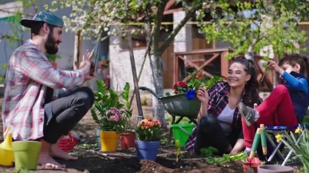 Счастливая атмосфера в саду молодая мама с маленьким ребенком сажают цветы и позируют перед камерой, в то время как папа фотографирует с помощью планшета — стоковое видео