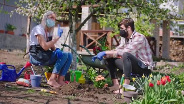 Glada unga par tillbringar dagen i trädgården de tar några blommor från krukorna och planterade i marken de stannar hemma i karantän bär skyddsmask Covid-19 pandemi — Stockvideo