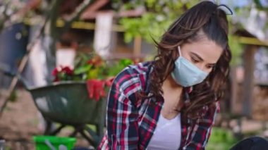 Bahçede çalışan genç ve güzel bir kadın. Su püskürtücü takmış çiçekler. Koruyucu maske takıyor ve Coronavirus 2019 karantinasında eldiven takıyor.