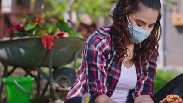 Привлекательная леди в защитной маске заботится о цветке в саду в то время как карантин она проводит время на свежем воздухе остаться дома пандемия коронавируса — стоковое видео