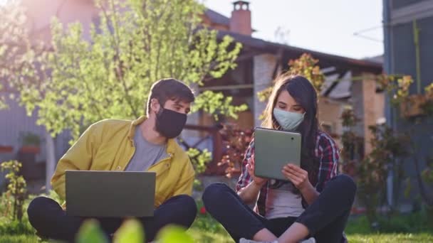 Pareja joven se quedan en casa en el jardín se sientan en el césped trabajando en línea mientras se quedan en cuarentena que usan máscaras protectoras usando tableta y computadora portátil para trabajar dama mostró algo a ella — Vídeos de Stock