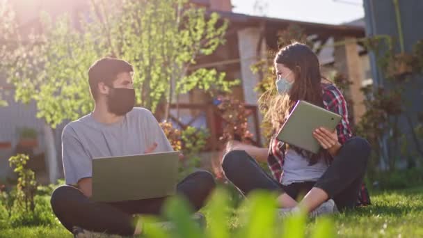 Красивая молодая женщина и ее харизматичный друг молодой парень беседуют, сидя в саду на траве и изучая онлайн вместе они носят защитную маску и остаются дома — стоковое видео