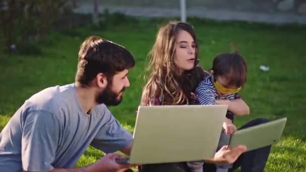 Mladí rodiče si užívají čas venku na zahradě, zatímco sedí na trávě a hrají si se svým synem, fotí se s otcem tabletu, který pracuje z notebooku zároveň. Výstřel na ARRI — Stock video
