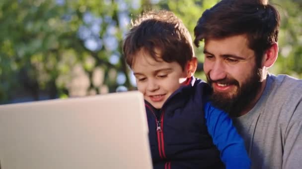 Piuttosto piccolo ragazzo con suo padre si divertono in giardino hanno una conversazione amichevole e utilizzando un computer portatile per giocare insieme un gioco. Girato su ARRI Alexa Mini — Video Stock