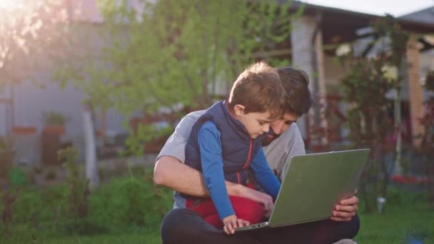 Une bonne journée pour un père et son joli garçon mignon ils s'assoient sur l'herbe dans le jardin et bavardent ensemble sur l'avenir ils utilisent l'ordinateur portable pour prendre des notes sur les rêves de l'enfant — Video