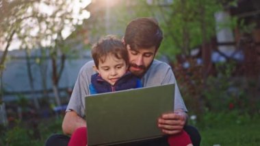 İyi vakit geçiren koca çeneli genç baba ve küçük oğlu bahçede eğleniyorlar. Dizüstü bilgisayarlarında oyun oynuyorlar ve çok heyecanlılar.