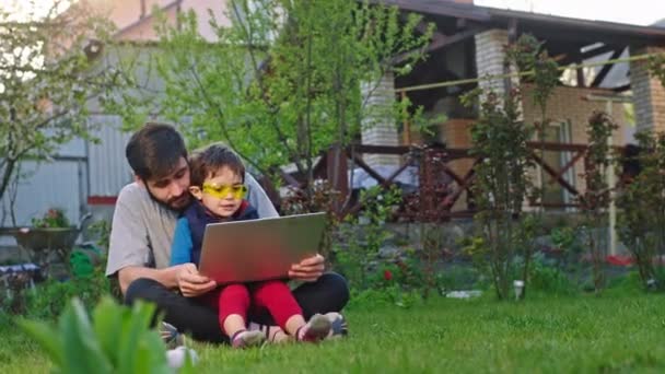 Em casa no jardim bom olhar bonito menino e seu pai se divertir juntos eles jogando juntos algum jogo de vídeo no laptop pequena criança vestindo óculos de sol engraçados — Vídeo de Stock
