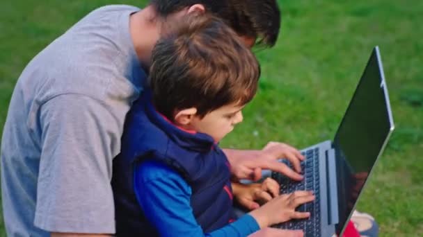 Wspaniały czas ojciec i mały syn w ogrodzie grając w jakąś grę wideo na laptopie cha razem mały chłopiec charyzmatyczny wyjaśnia coś swojemu tacie — Wideo stockowe