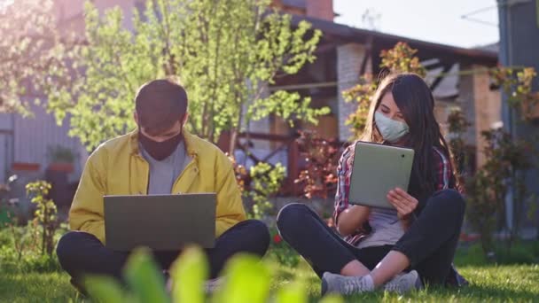 Солнечный день дома в саду привлекательные леди и мужчина сидит в траве в то время как Qua они учатся из дома с помощью планшета и ноутбука они носят защитную маску новые Covid-19 пандемии — стоковое видео