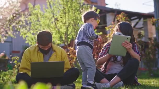 Hemma mamma och pappa arbetar i trädgården medan du sätter dig på gräset de använder laptop och surfplatta för att arbeta online bär skyddsmask deras lilla son leker runt dem — Stockvideo