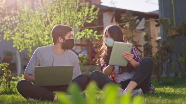 В солнечный день пара остаться дома и учиться вместе с помощью гаджета они находятся в карантине нового Ковид-19 они носят защитную маску, сидя в саду на траве — стоковое видео