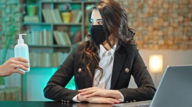 Ofisteki kameranın önündeki portre. Koruyucu maskeli, laptopunda çalışan güzel bir kadın. Bir el ona dezenfekte edilmiş jel veriyor.