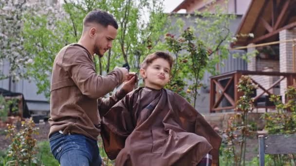 Barbeiro homem ter um tempo divertido com um menino pequeno fazer um corte de cabelo carismático menino pequeno fazer um rosto engraçado e se sentindo relaxado — Vídeo de Stock