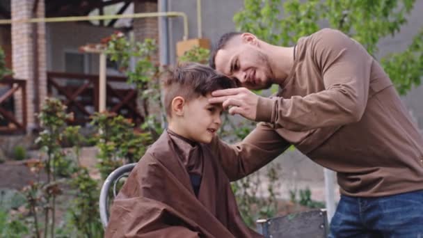 Carismático niño pequeño tiene un corte de pelo en el jardín se para y hacer una linda cara sonriente barbero hombre tener una charla con el niño pequeño — Vídeos de Stock