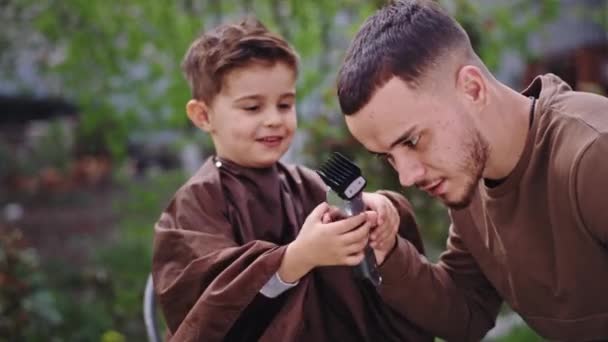 Tempo de corte de cabelo no jardim menino carismático apreciando o tempo com o barbeiro que ele dá ao menino o clipper eletrônico para tentar fazer um corte de cabelo no barbeiro — Vídeo de Stock