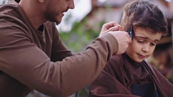 Vor der Kamera charismatische kleine süße Junge haben eine Frisur im Garten Friseur Mann arbeitet konzentriert mit einer elektronischen Haarschneidemaschine — Stockvideo