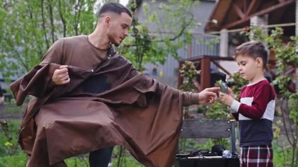 Anak laki-laki kecil yang lucu mencoba untuk membuat potong rambut untuk kakaknya di kebun ia mengambil gunting elektronik dan mulai bekerja — Stok Video