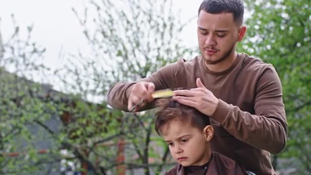 W ogrodzie na zewnątrz fryzjer zrobić fryzurę do ładny i smutny mały chłopiec skoncentrowany obciął włosy nożyczkami — Wideo stockowe
