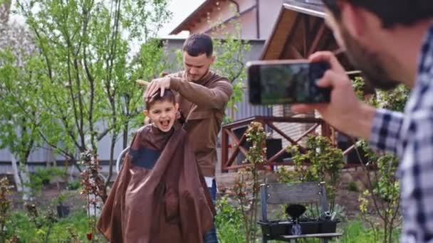 庭で大きな面白いかわいい男の子を笑顔誰かが彼が満足している小さな男の子の写真を撮っている間床屋はヘアカットを作る — ストック動画