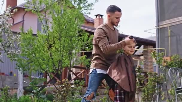 Leuke tijd voor een grote broer en zijn kleine broer maken een kapsel in de tuin met een elektronische tondeuse voor de camera — Stockvideo