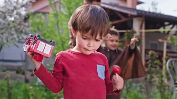 Cute small boy in the garden with a lollipop in front of the camera make a funny face while holding a car background big brother make a haircut — Stock Video