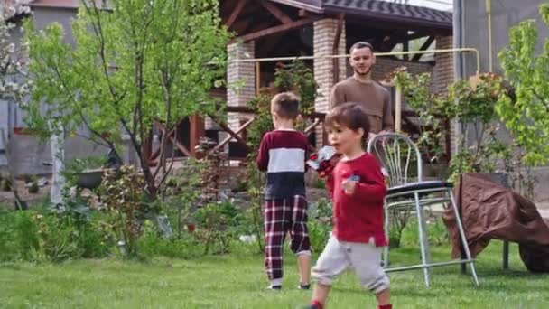 An einem sonnigen Tag im Garten amüsieren sich zwei kleine Kinder mit ihrem großen Teenager-Bruder, den sie gemeinsam auf dem Rasen spielen. — Stockvideo