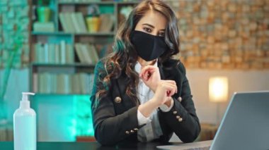 Koruyucu maskeli genç bir kadın ofis çalışanı laptopta çalışmaya başlamadan önce ellerini dezenfekte etmesini ve Coronavirus 2019 konsepti üzerinde çalışmaya devam etmesini önerdi.