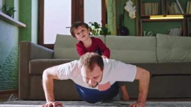 Babasıyla birlikte gün batımında oturma odasında spor yapan komik bir çocuk. Babası sırtında küçük bir çocukla şınav çekiyor. ARRI Alexa Mini 'de çekilen