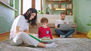 Evde oğluyla kitap okuyan genç bir anne arka planda oturan baba laptopuyla evde çalışırken yerde oturuyor.
