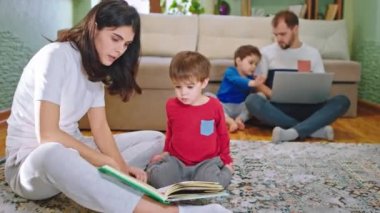 Geniş bir aile bütün gününü evde geçiriyor. Anne, oğluyla bir hikaye okuyor. Diğer çocukla birlikte dizüstü bilgisayardan bir şeyler yapıyor.