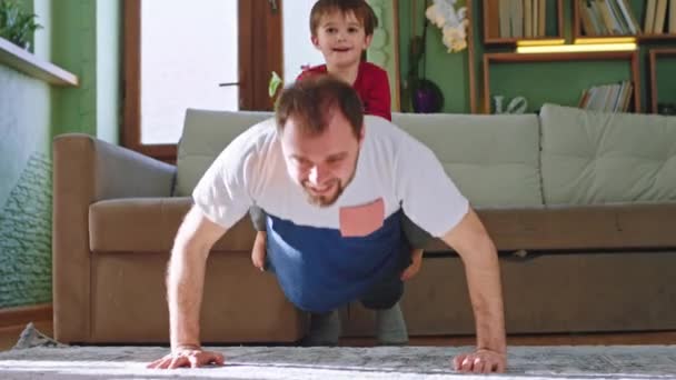 Buen momento en casa padre con su pequeño niño haciendo deporte en la alfombra en la sala de estar niño sentado en su padre de vuelta mientras que el padre haciendo flexiones — Vídeo de stock