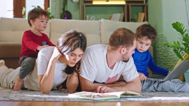 Auf dem Boden liegen glückliche Mütter und Väter, die Zeit mit ihren beiden Kindern verbringen und die Geschichte aus dem Buch vorlesen und am Laptop spielen. — Stockvideo