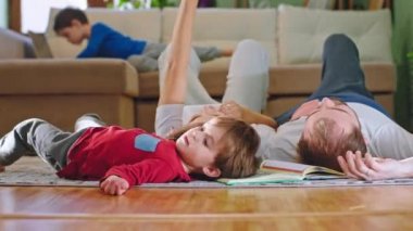 Küçük şirin bir çocuk ve ebeveynleri yerde yatarken birlikte vakit geçiriyorlar. Diğer koca oğlan kanepede oturuyor ve dizüstü bilgisayarında oyun oynuyor.