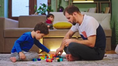 Baba ve büyük oğlu oturma odasında yerde otururken birlikte oynuyorlar diğer küçük çocuklar kanepede televizyon izliyorlar. 4k