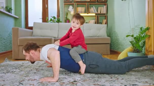 Deporte tiempo en casa en la sala de estar papá con su pequeño hijo lindo haciendo flexiones juntos mientras que el niño pequeño está en su espalda — Vídeo de stock