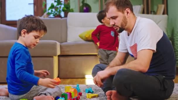 Glücklicher Vater mit seinen beiden Kindern, die Zeit zu Hause verbringen, während ein Kind fernsieht, während er mit seinem Sohn unten auf dem Boden im Wohnzimmer spielt — Stockvideo