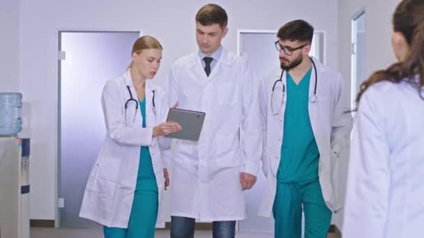 En el hospital el médico principal en el pasillo caminando al lado de su médico universitario y enfermeras y hablando de algo de fondo en la silla paciente tener una charla con su médico de familia — Vídeo de stock