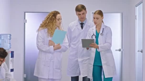 Главный врач взрослый мужчина и его коллега молодые две женщины врачи, проходящие через больничный коридор и беседующие они анализируют с планшета некоторые результаты тестов своего пациента — стоковое видео