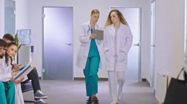 Büyük hastane koridorunda iki genç doktor ellerinde dijital tabletle kameranın önünde yürüyorlar ve hasta sonuçlarını analiz ediyorlar.