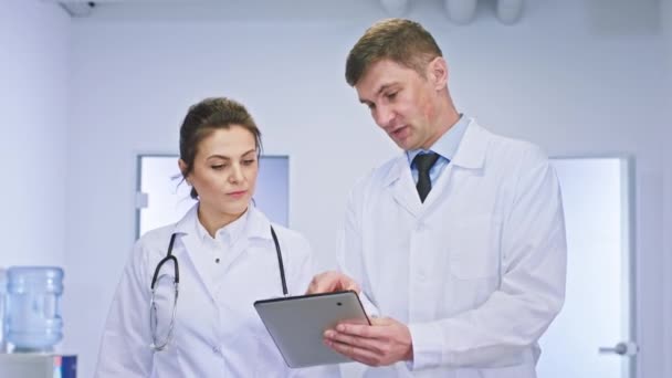 En un pasillo luminoso cerca de la cámara, el hombre y la mujer médicos discuten el diagnóstico del paciente con una tableta digital — Vídeo de stock