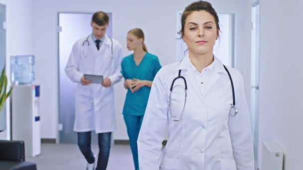 Kameranın önünde modern bir hastane koridorunda kameranın önünde yürüyen kadın doktorun arka planında doktor ve hemşire asistanı tanıyı analiz ediyor ve tartışıyor. — Stok video