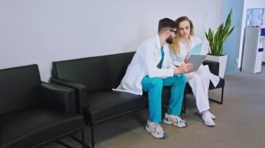 Kadın ve erkek doktorlar hastanenin modern koridorunda yürüyorlar. Sandalyedeki diğer hemşireler ve doktorlar da tartışıyorlar.