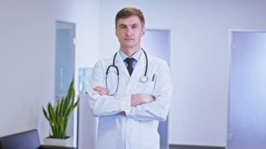 Kameranın önünde ciddi bir suratı olan doktor portresinin üzerinde steteskop var. Hastane koridorunda duruyor.