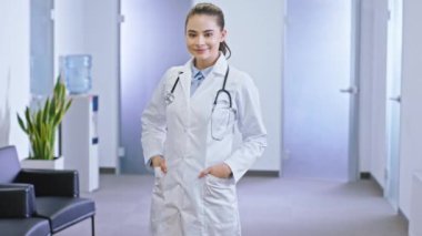 Kameranın önünde duran karizmatik gülümsemeli bayan doktor doğrudan kameraya bakıyor hastane koridorunda mutlu bir yüzü var.
