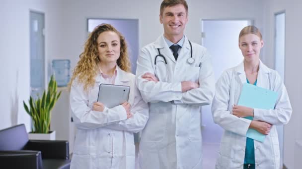 Портрет улыбающихся больших врачей перед камерой в центре больничного коридора дамы-врачи с цифровыми планшетами и картами — стоковое видео