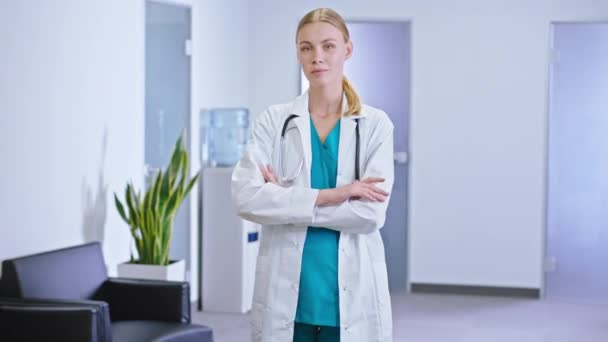 Hermosa joven enfermera mirando directamente a la cámara en un moderno pasillo del hospital ella sonriendo lindo usando el estetoscopio — Vídeos de Stock