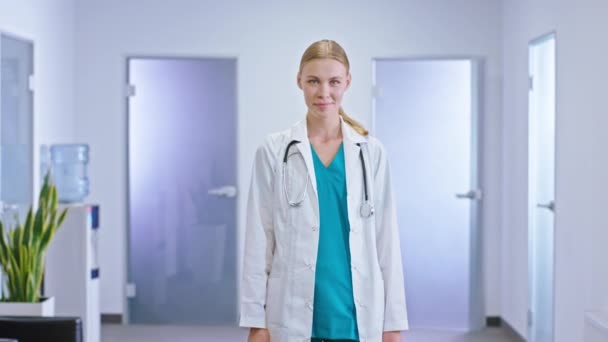 Счастливая молодая женщина семейный врач смотрит прямо в камеру и иметь хорошее настроение в коридоре больницы — стоковое видео